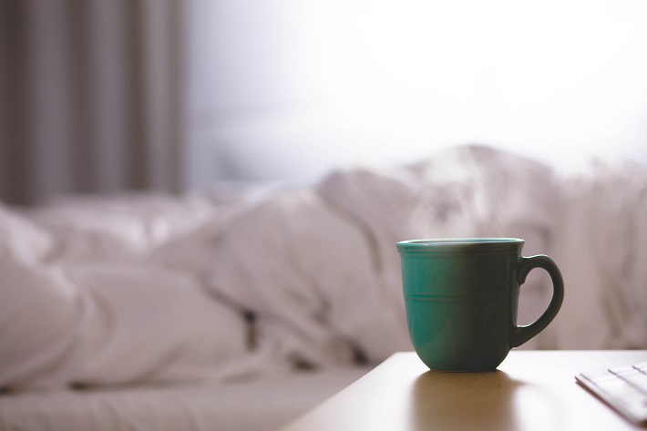 Steaming cup of tea on a bedside table next to someone bundled up in a duvet